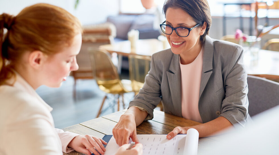 Zwei Frauen sitzen am Tisch, die eine Frau zeigt auf eine Tabelle