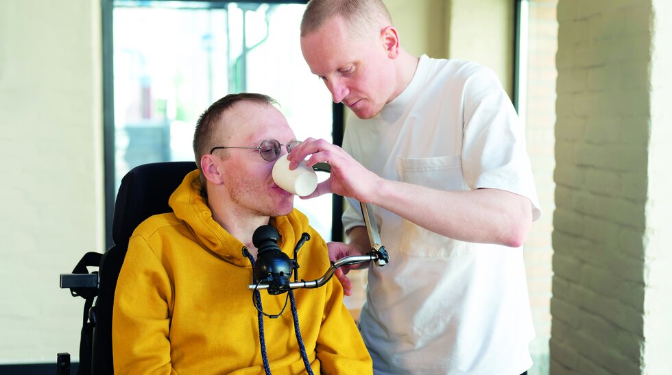 Ein Pfleger hilft einen Mann beim Trinken, der im Rollstuhl sitzt