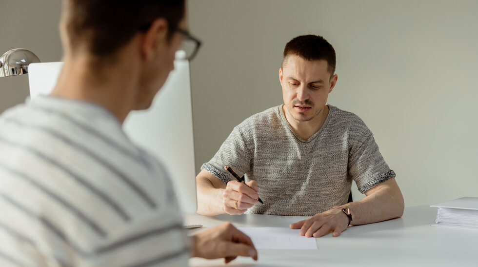 Zwei Männer sitzen sich am Tisch gegenüber und gucken auf ein Blatt Papier.