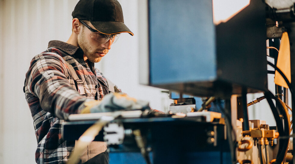 Umschulung Fachpraktiker/-in Metalltechnik Ein Mann mit Kappe steht an einem Gerät und arbeitet