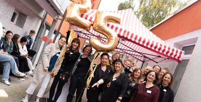 Mitarbeiterinnen von Der Wendepunkt stehen vor einem Pavillon und halten Luftballons mit 2 und 5 in die Höhe.