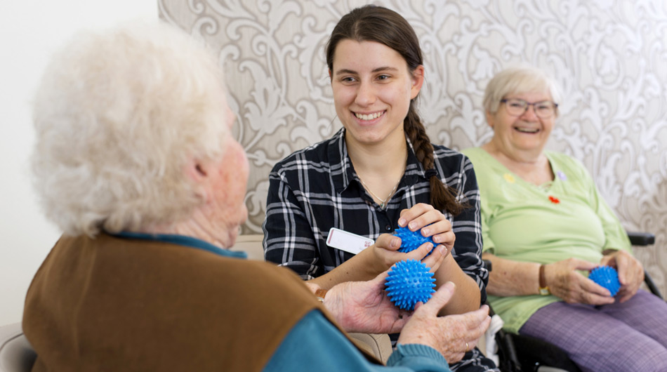 Eine Schülerin der Ausbildung "Sozialassistent" und zwei Seniorinnen machen Handübungen mit kleinen Bällen.