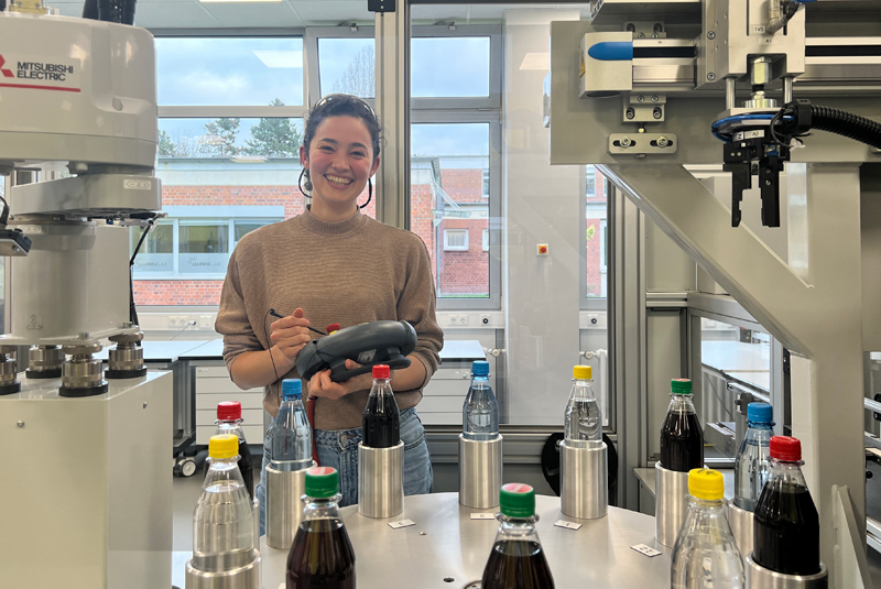 Nadja Graschi Maschine Nadja steuert einen Roboter, sie hat eine Schutzbrille an. Vorne stehen Flaschen.