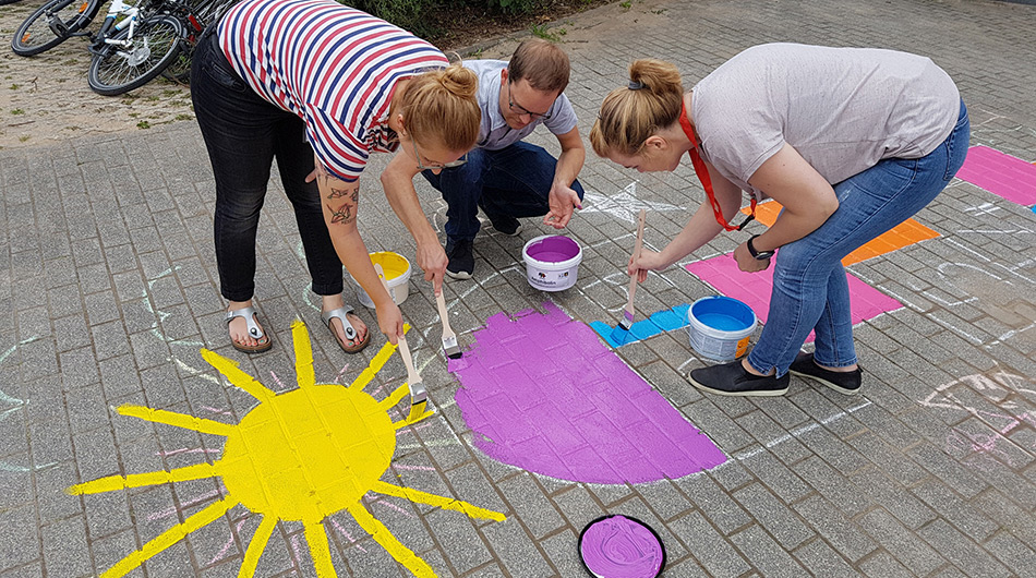 Zwei Frauen und ein Mann malen während ihres Unternehmensengagements bunte Symbole auf den Boden.