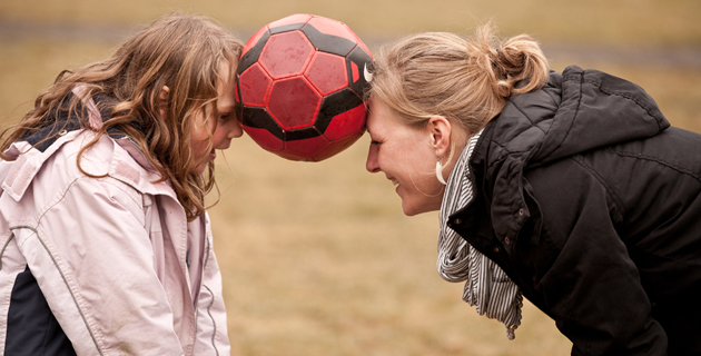 Über uns: Eine Erzieherin und ein junges Mädchen halten einen Fußball zwischen ihren Stirnen.